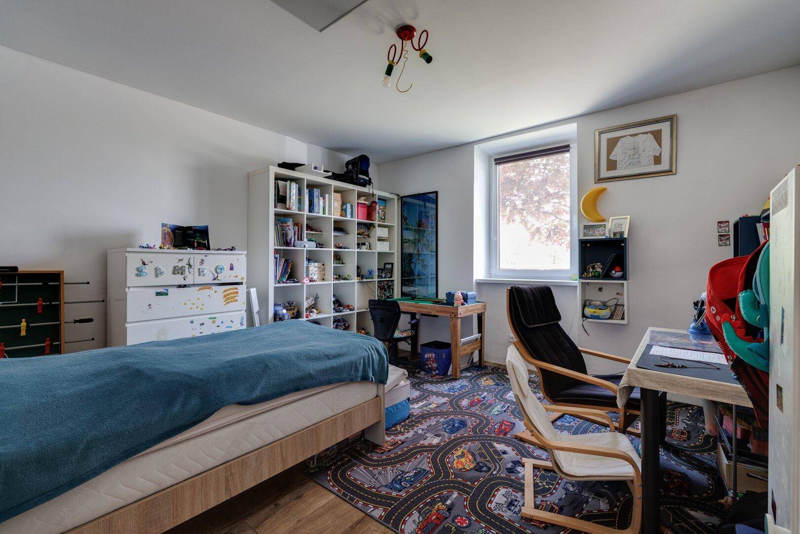 Kinderzimmer mit Einzelbett, Schreibtisch, Bücherregal und einem spielerischen Teppich.