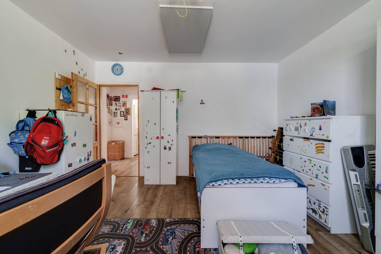Helles Kinderzimmer mit zwei Betten, Kommode und einem bunten Spielteppich.
