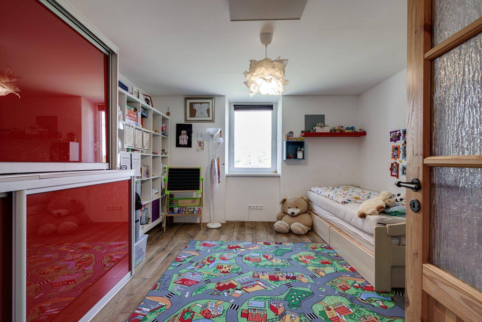 Kinderzimmer mit Einzelbett, Schreibtisch, Bücherregal und einem auffälligen roten Spiegelschrank.