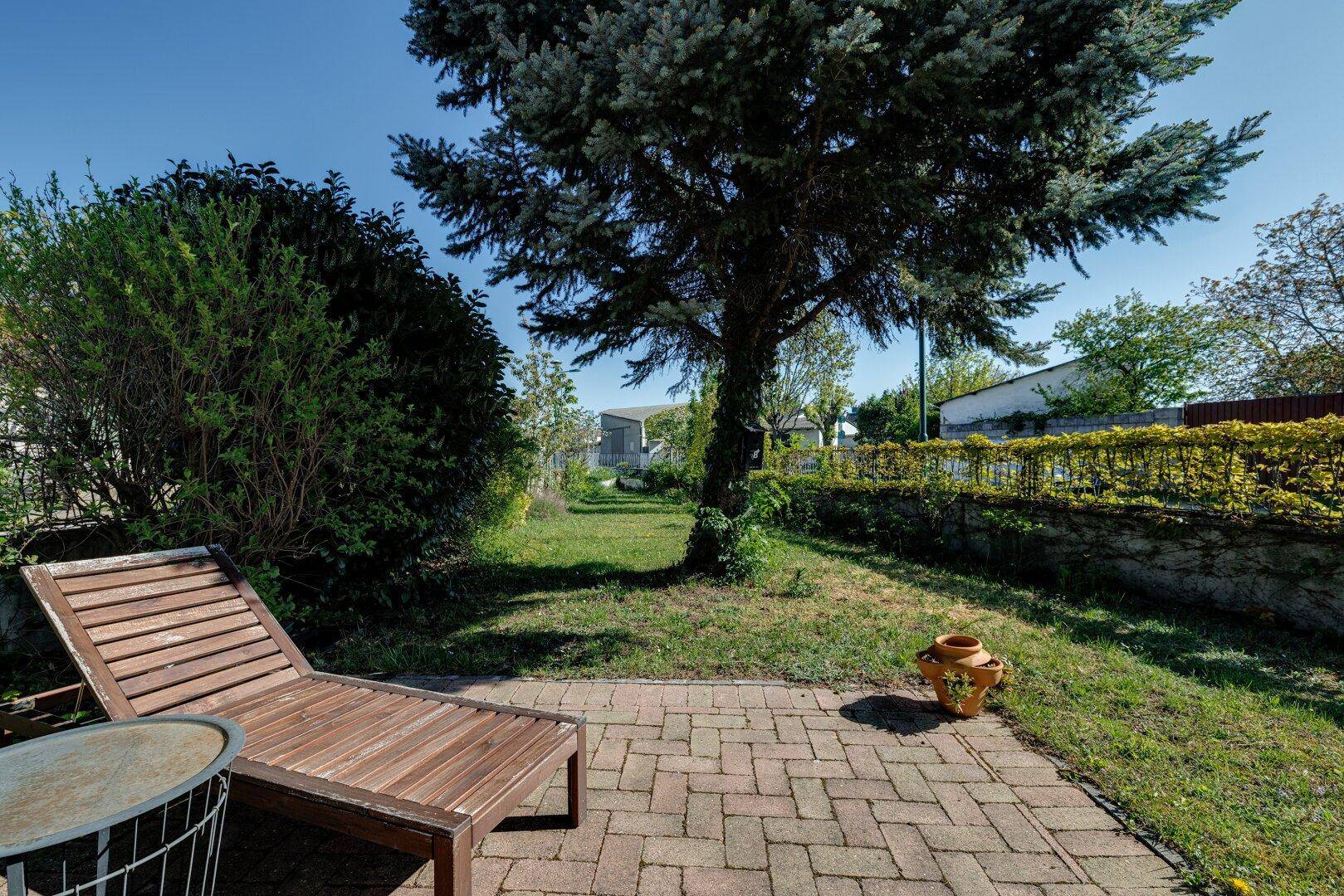 Gepflegter Garten mit Rasen, Baumbestand und gemütlicher Liegefläche auf der Terrasse.