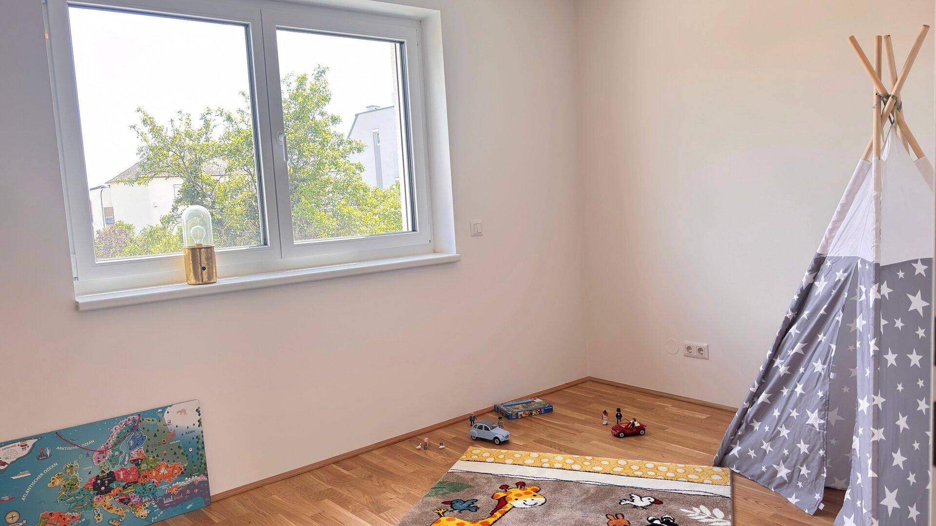 Kinderzimmer mit Holzboden, großem Fenster und einem Spielteppich sowie einem Tipi-Zelt.