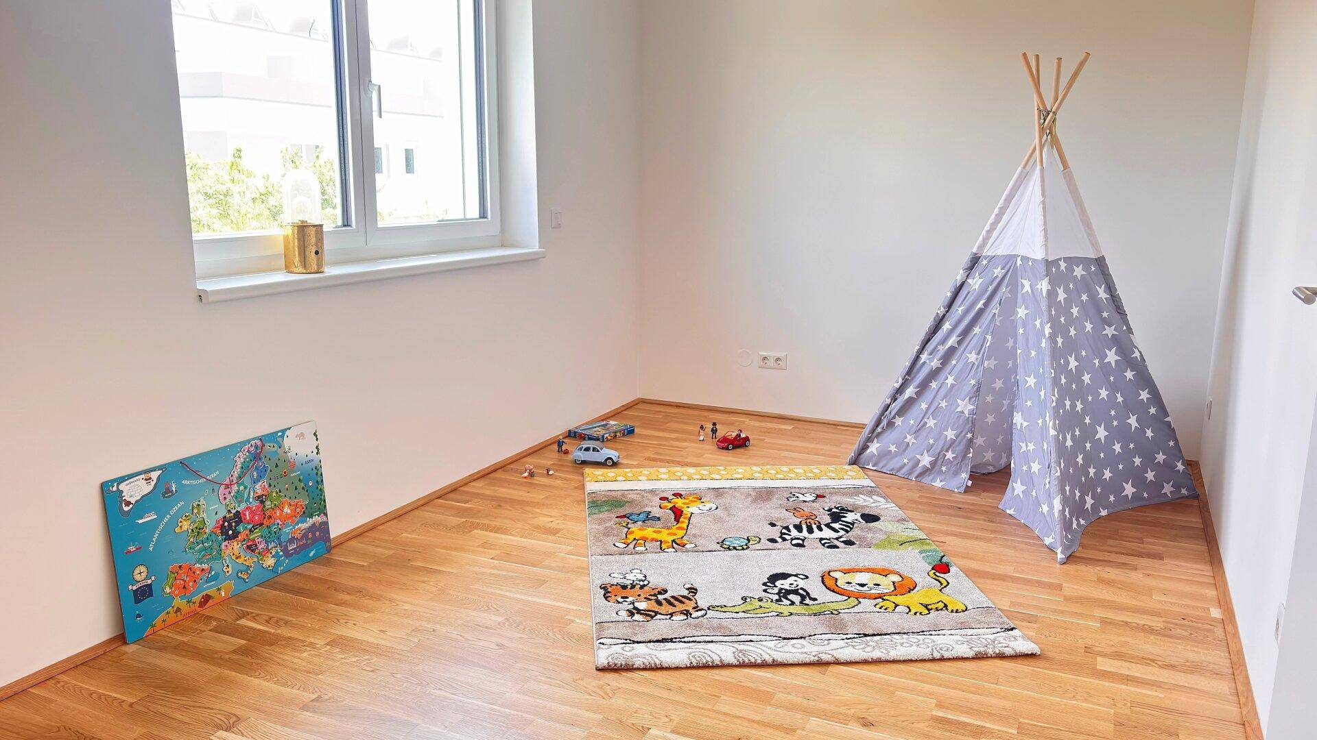 Helles Kinderzimmer mit Parkettboden, großem Fenster und einem bunten Spielteppich.