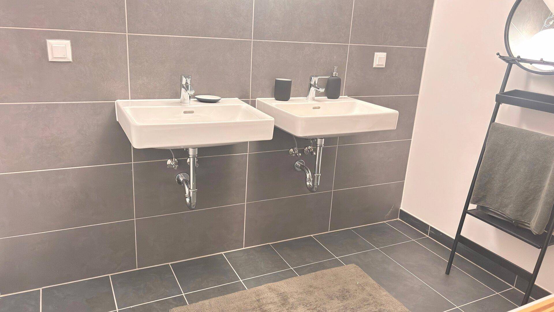 Modernes Badezimmer mit zwei Waschbecken, großen grauen Fliesen und einem Spiegel.