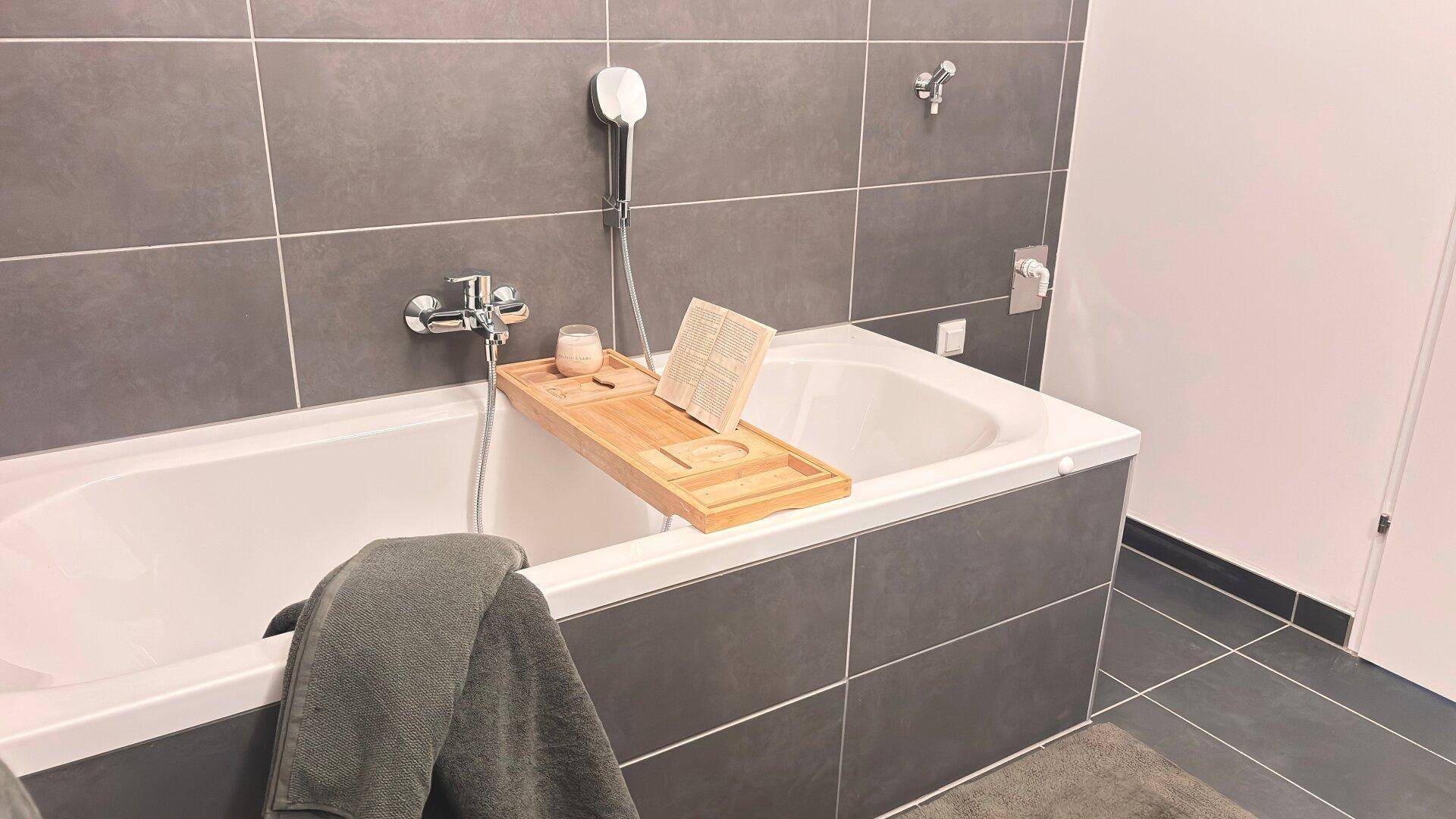 Modernes Badezimmer mit Badewanne, grauen Fliesen und einer Ablage für Badutensilien.