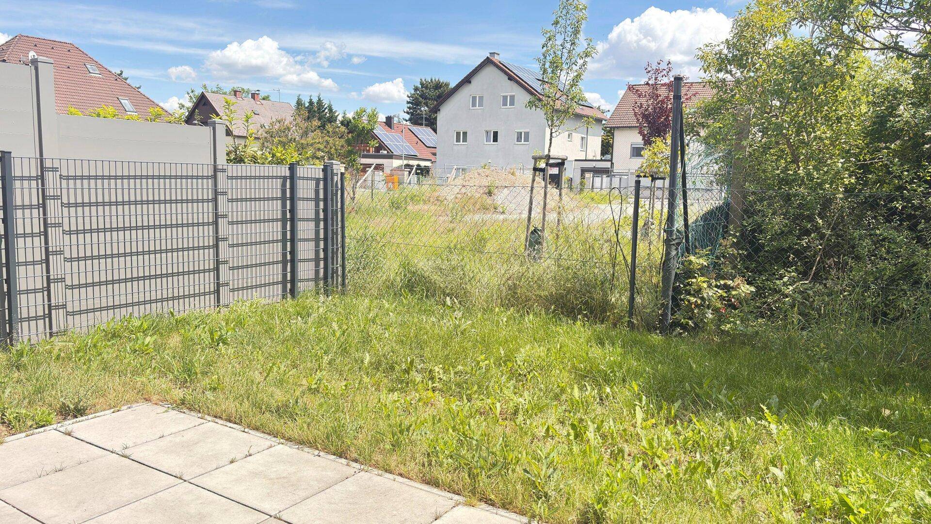 Ungepflegte Gartenfläche mit Zaun und Blick auf benachbarte Wohnhäuser unter blauem Himmel.
