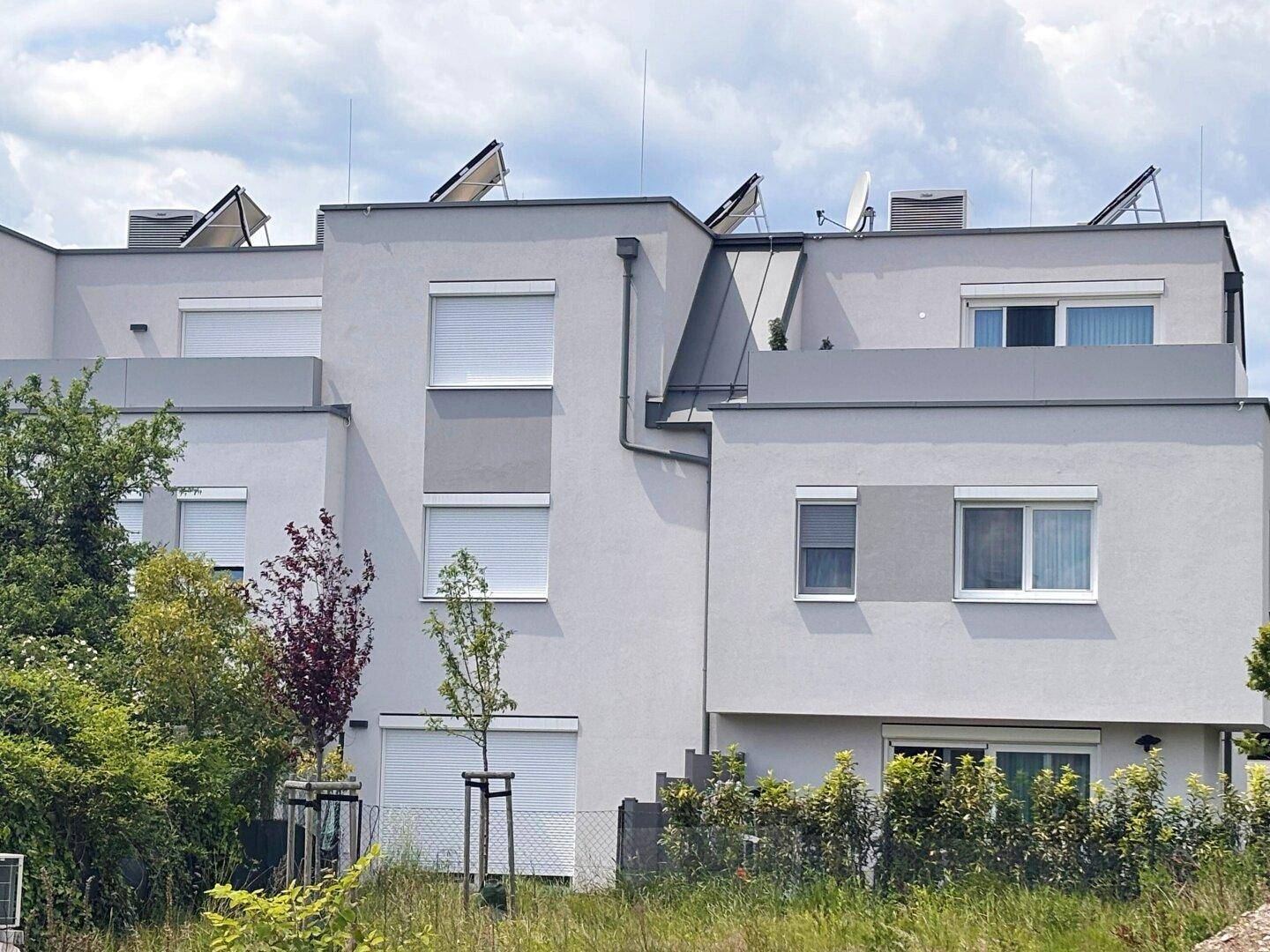 Moderne Gebäudefassade mit Flachdach, Rollläden und Solaranlagen auf dem Dach.