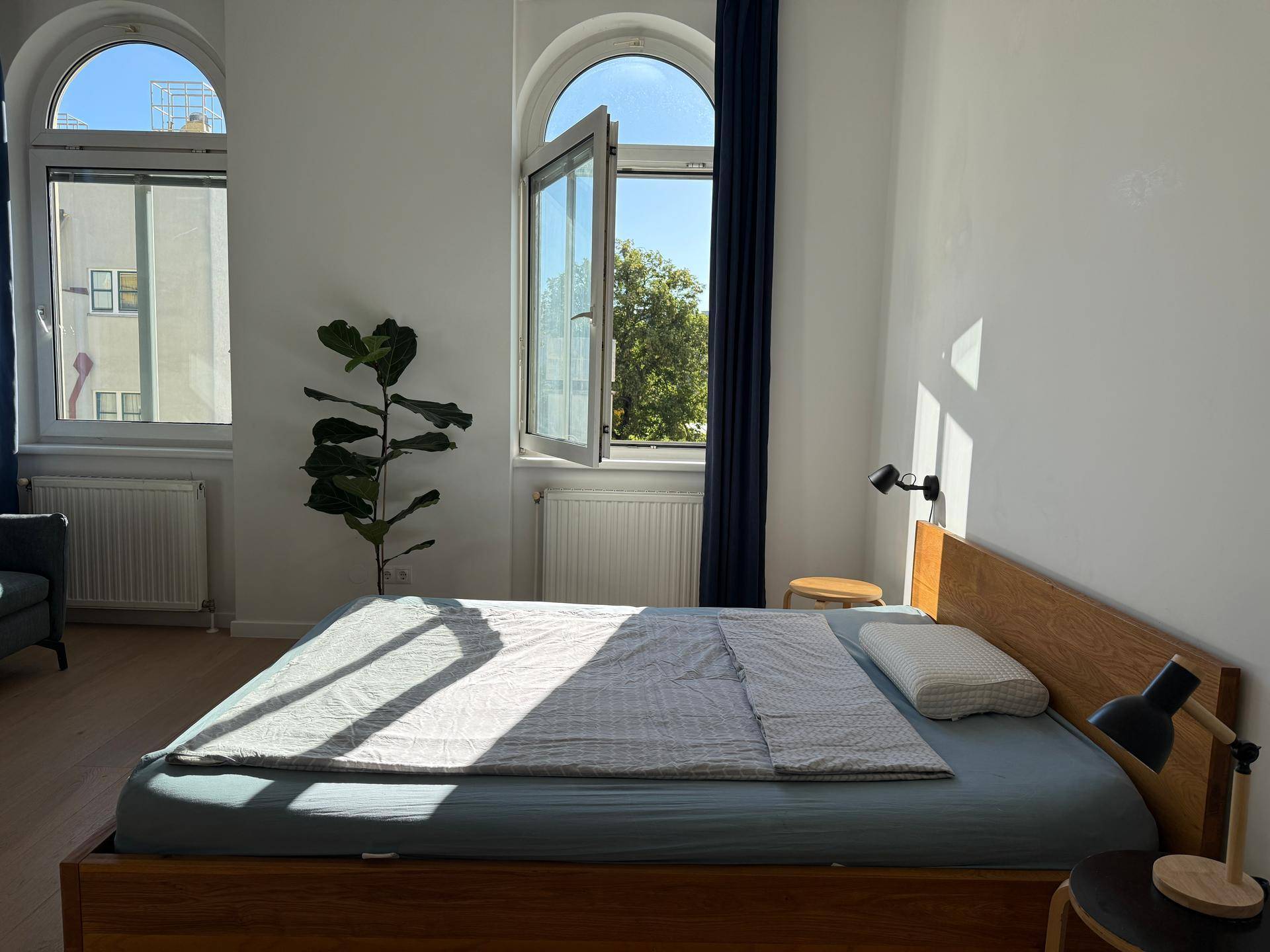 Sonnendurchflutetes Studio-Apartment mit Holzbett und geöffnetem Rundbogenfenster, das einen Blick ins Grüne bietet.
