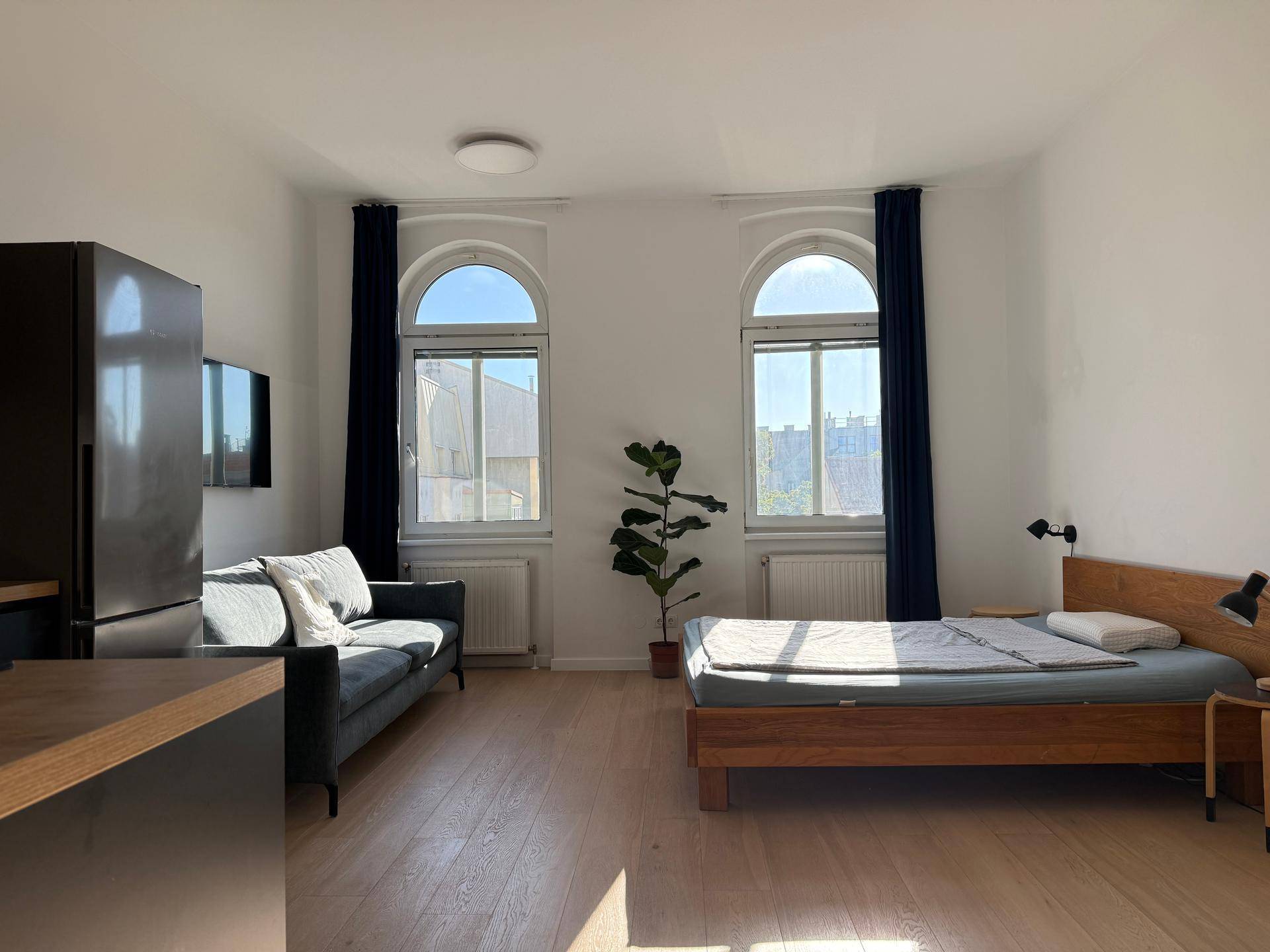 Geräumiges Studio-Apartment mit Holzbett, Sofa und einem modernen Kühlschrank, beleuchtet von Tageslicht.