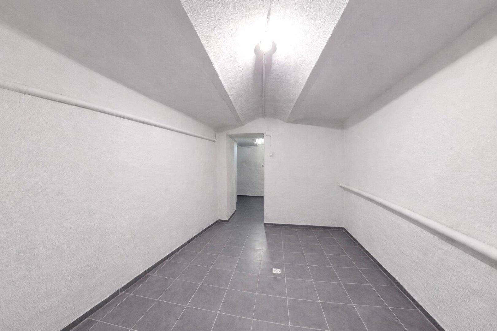 Kellerraum mit weiß gestrichenen Wänden, grauen Fliesen und sichtbaren Rohren an der Decke.