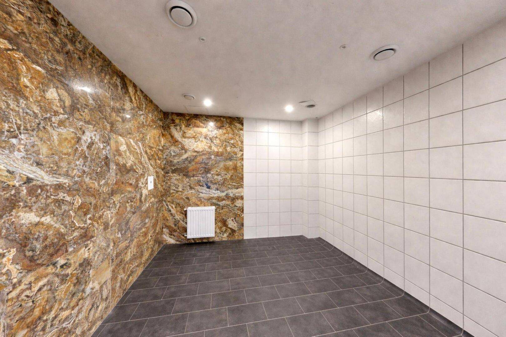 Raum mit einer Wand aus Naturstein und drei Wänden mit weißen Fliesen, sowie dunklem Fliesenboden.