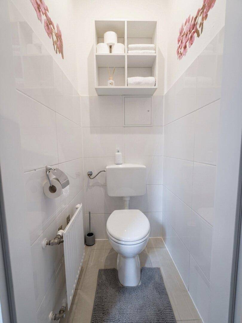 Separates WC mit Wandtoilette, kleinem Heizkörper und praktischem Regal.