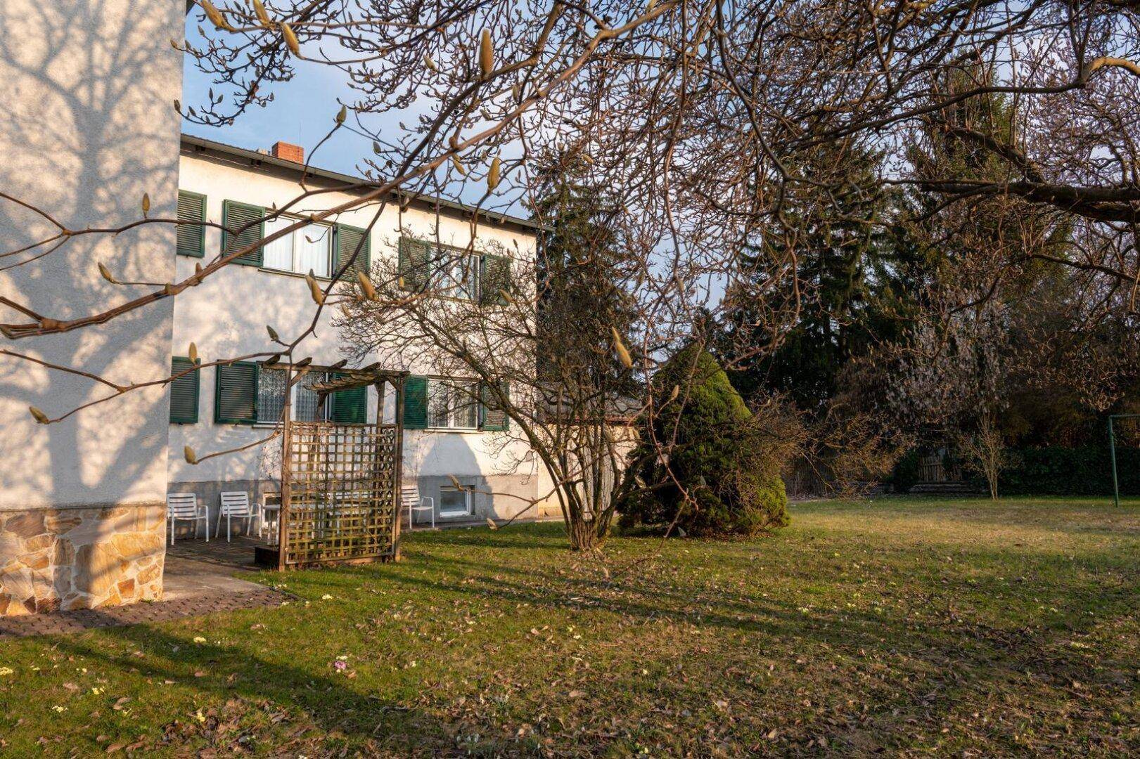 Weitläufiger Garten mit Rasen, Bäumen und einem kleinen Sitzbereich neben dem Haus.