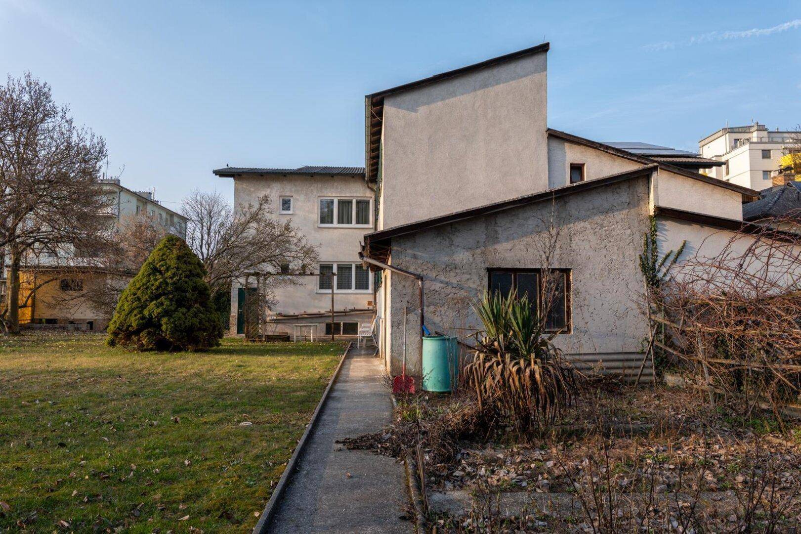 Rückansicht des Hauses mit einem schmalen Weg und einem ungepflegten Gartenbereich.