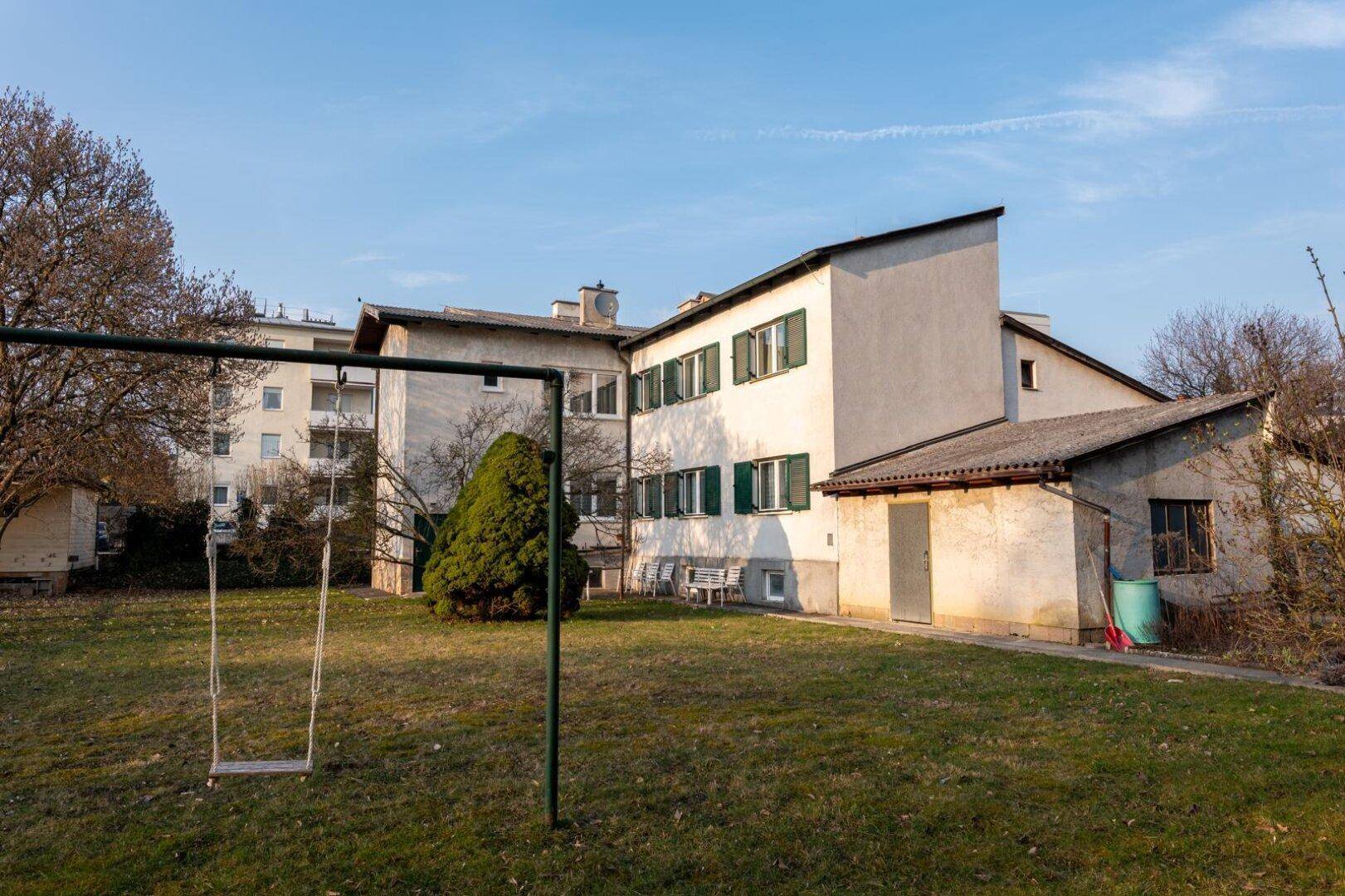 Großer Garten mit Schaukel und Blick auf die Rückseite des Mehrfamilienhauses.