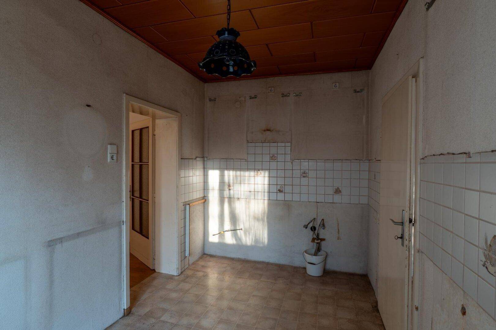 Leere Küche mit alten Fliesen, Holzdecke und sichtbaren Anschlüssen für eine Spüle.