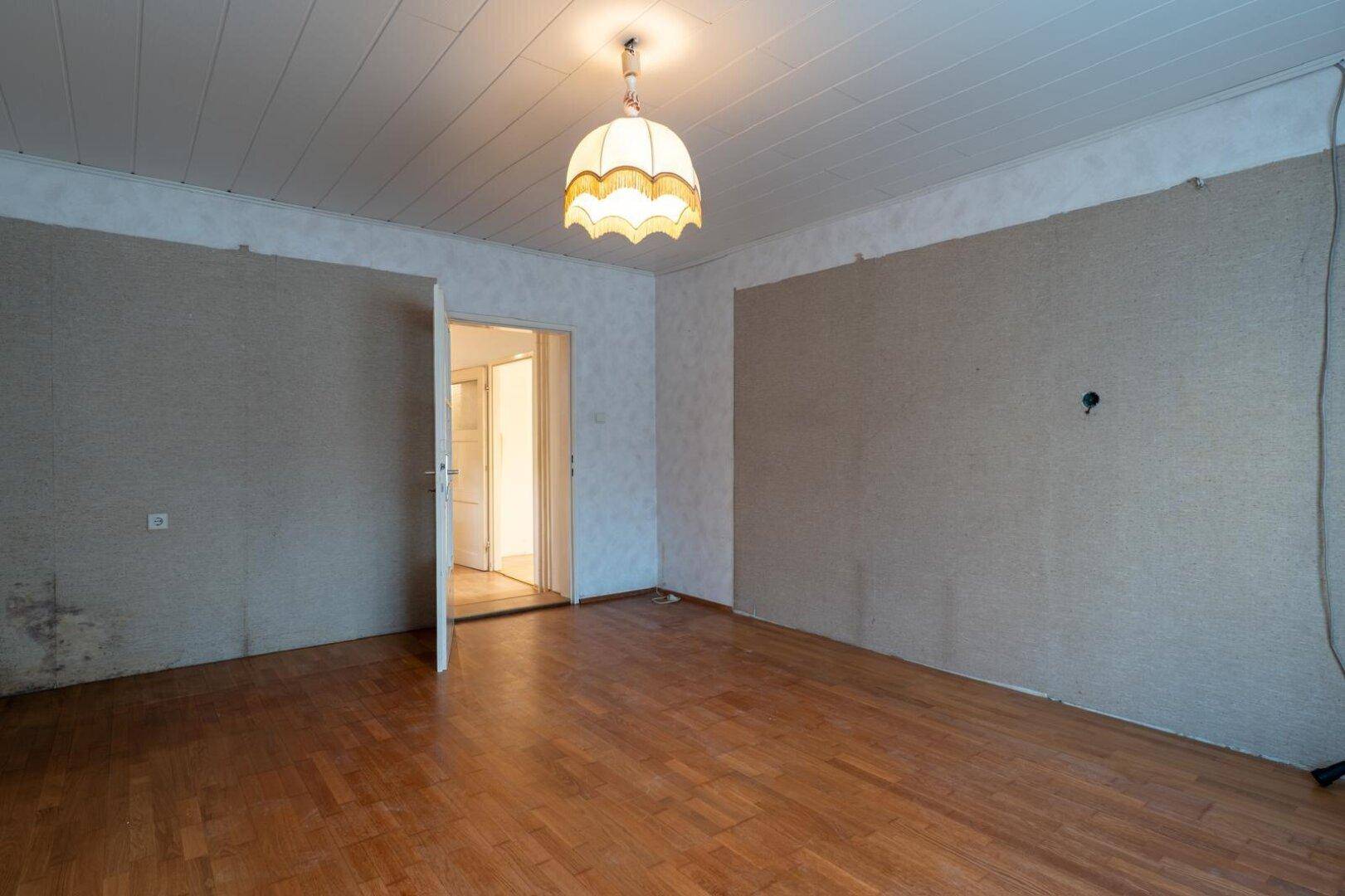 Leeres Zimmer mit Holzboden, weißer Decke und einer offenen Tür zu einem weiteren Raum.