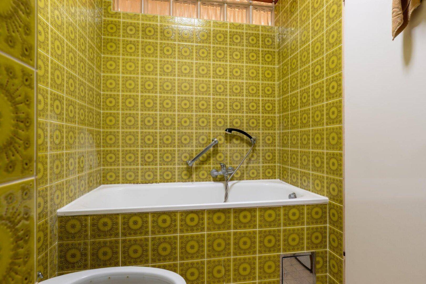 Badezimmer mit gelb-grünen Fliesen und eingebauter Badewanne unter einem Glasbausteinfenster.