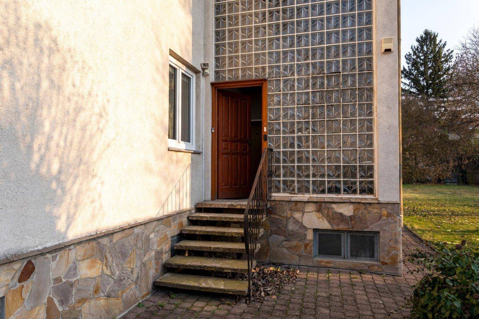 Eingangsbereich des Hauses mit Steintreppe, Holztür und großer Glasbausteinwand.