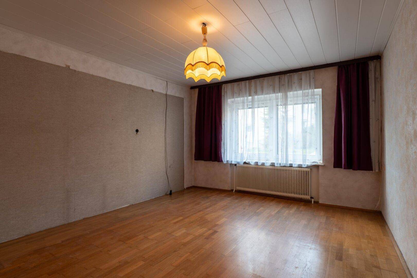 Helles Zimmer mit Holzboden, Heizkörper und Fenster mit Vorhängen, bereit zur Renovierung.