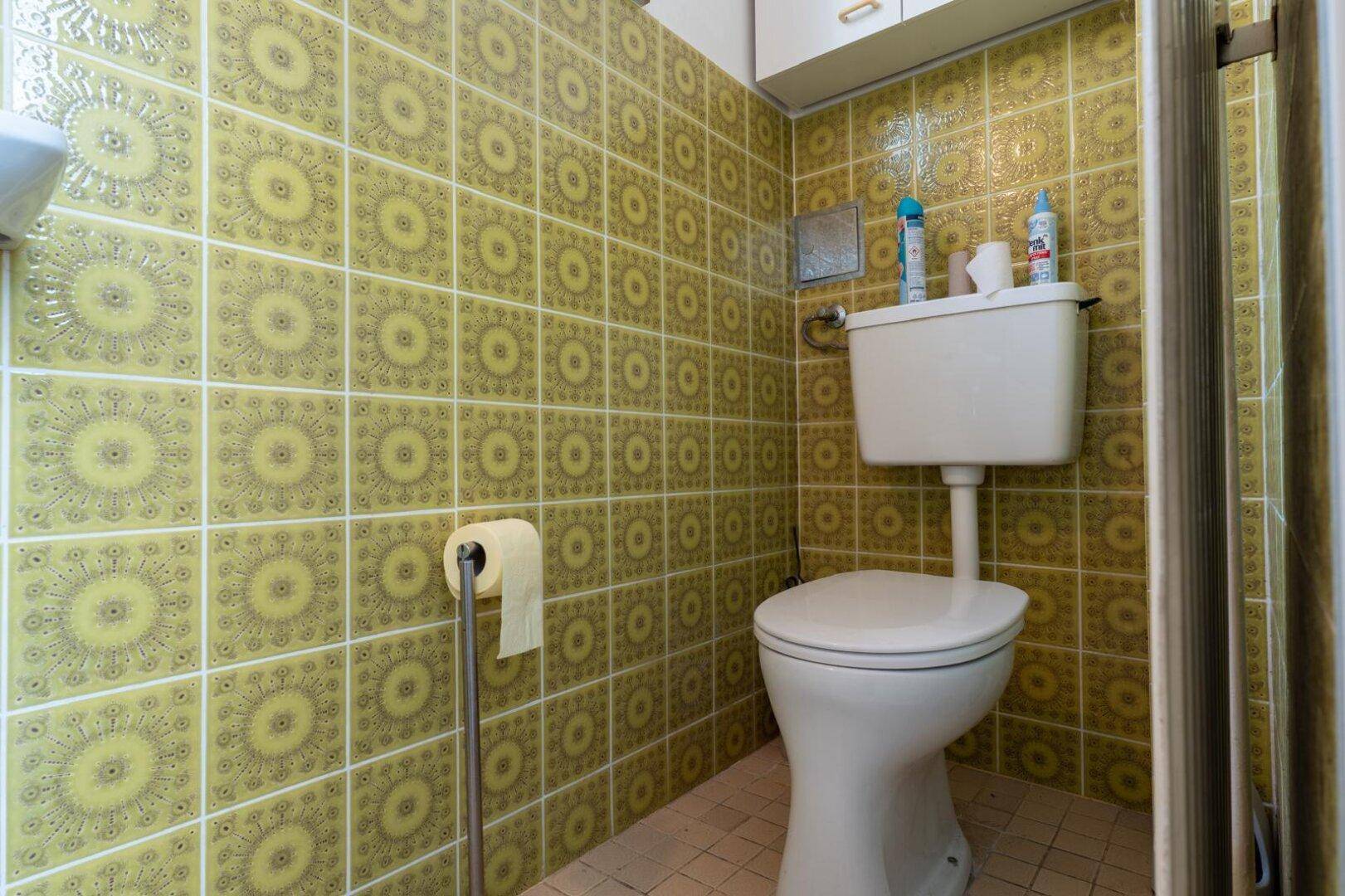 Toilette mit gelb-grünen Fliesen und einem Spülkasten über dem WC.