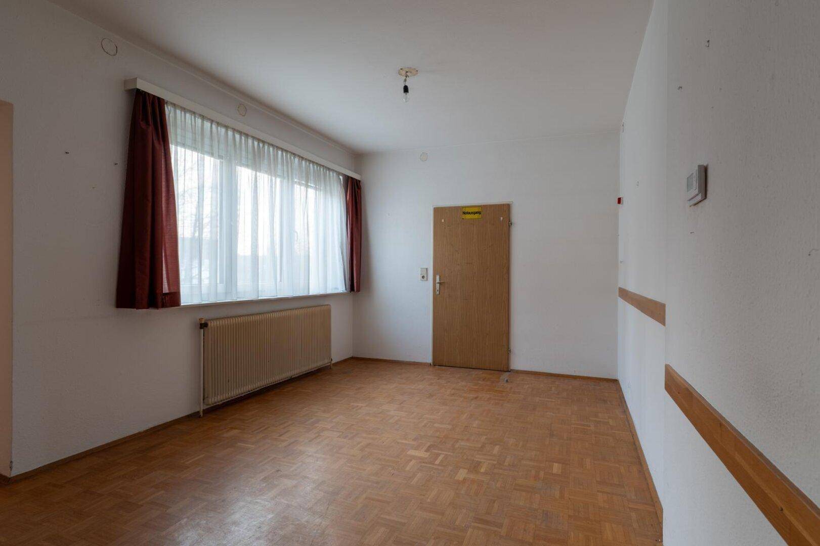Helles Zimmer mit Parkettboden, großem Fenster und einer Holztür.
