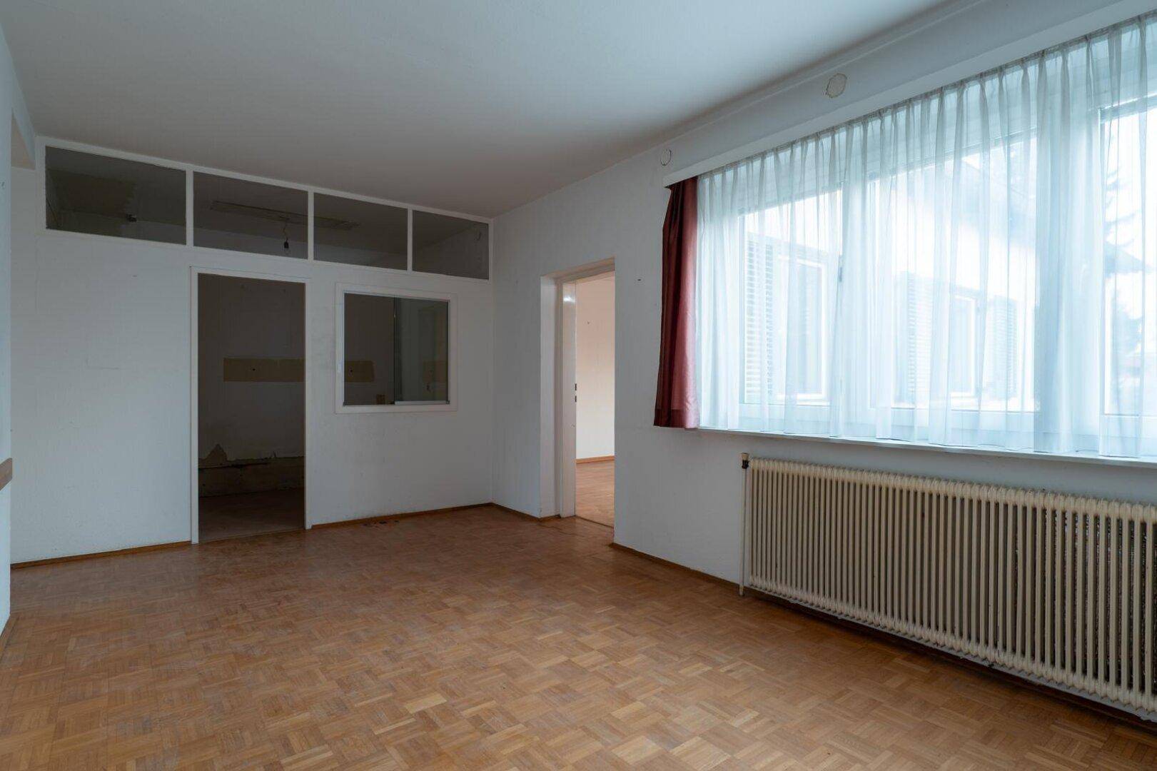 Geräumiges Wohnzimmer mit Parkettboden, großen Fenstern und Blick in einen weiteren Raum.
