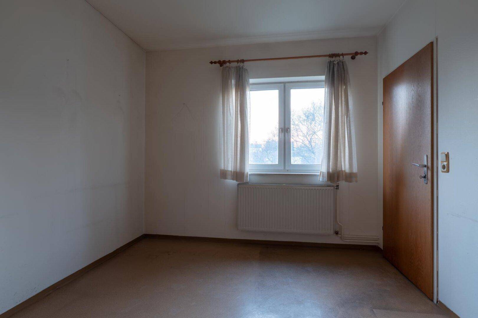 Helles Schlafzimmer mit Fenster, Heizkörper und einfacher Holztür.
