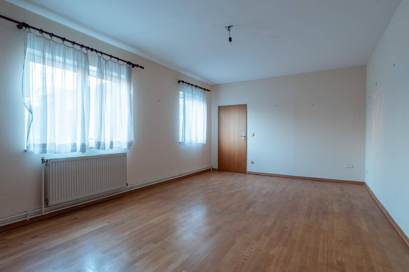 Geräumiges Zimmer mit Parkettboden, zwei Fenstern und einer Holztür.