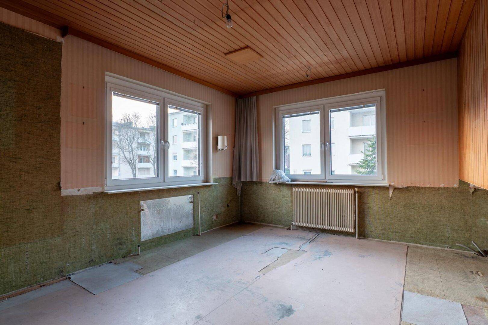 Geräumiges Zimmer mit Holzdecke, zwei Fenstern und abgenutzter Tapete, bereit zur Renovierung.