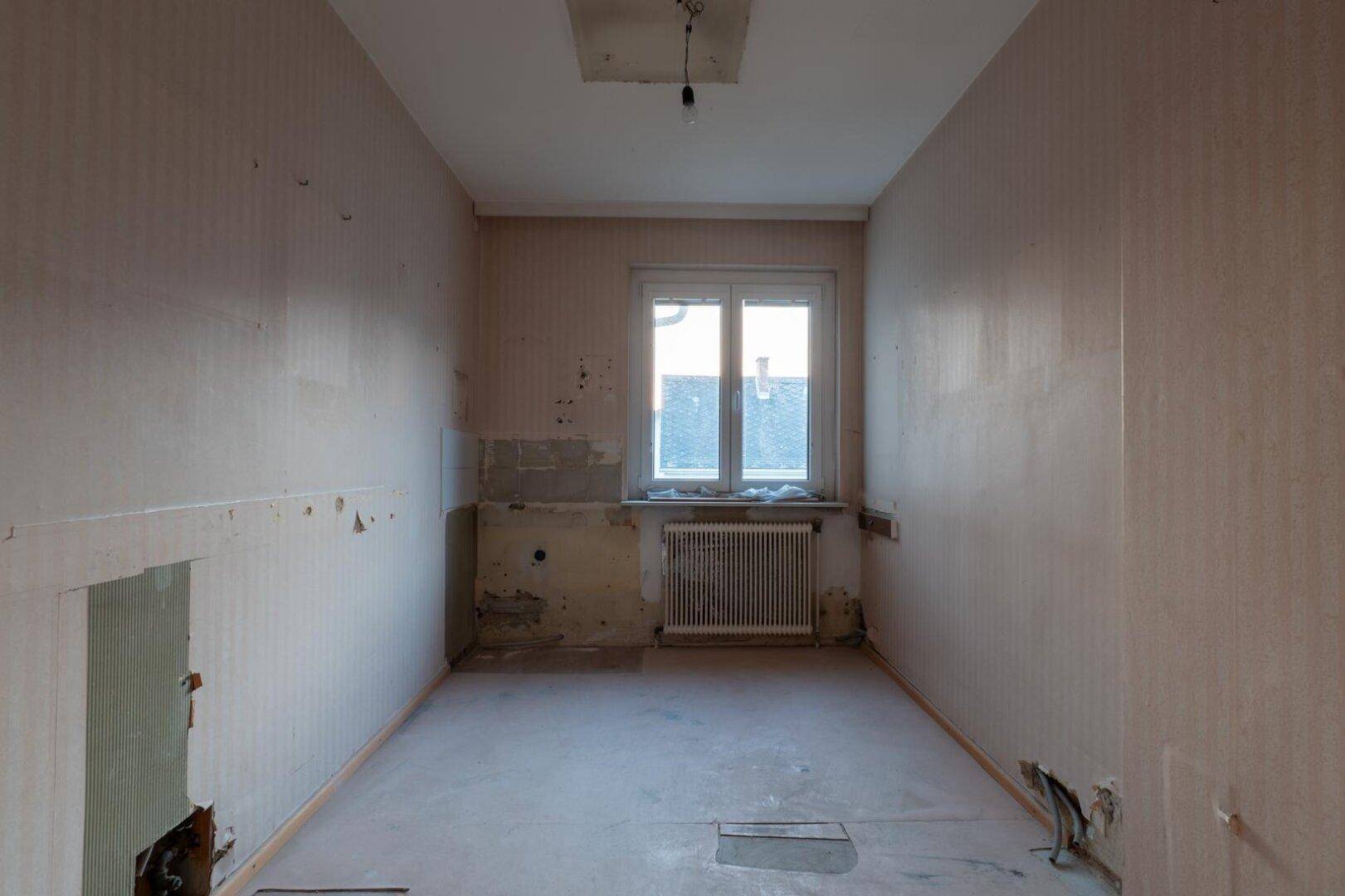 Leere Küche mit Fenster, Heizkörper und abgenutzten Tapeten, bereit zur Renovierung.