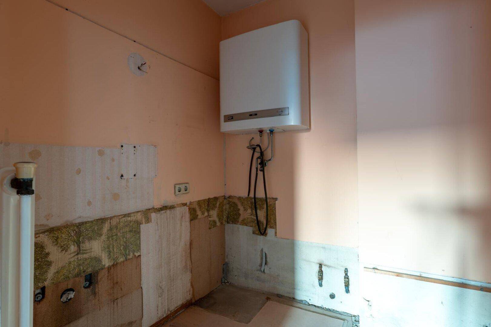 Küchenbereich mit Warmwasserbereiter, abgenutzter Tapete und sichtbaren Anschlüssen.