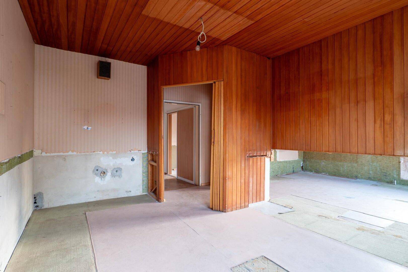 Innenraum mit umfangreicher Holzvertäfelung und Zugang zu weiteren Zimmern, renovierungsbedürftig.