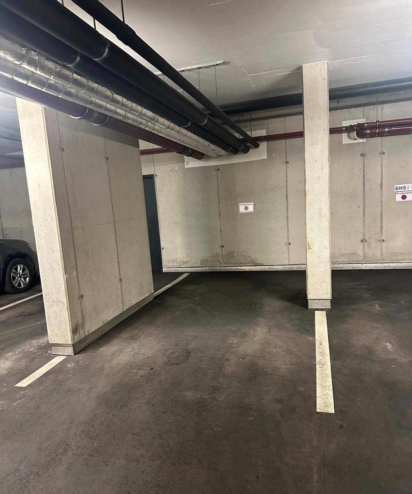 Innenansicht einer Tiefgarage mit Betonwänden, Säulen und sichtbaren Versorgungsleitungen an der Decke.