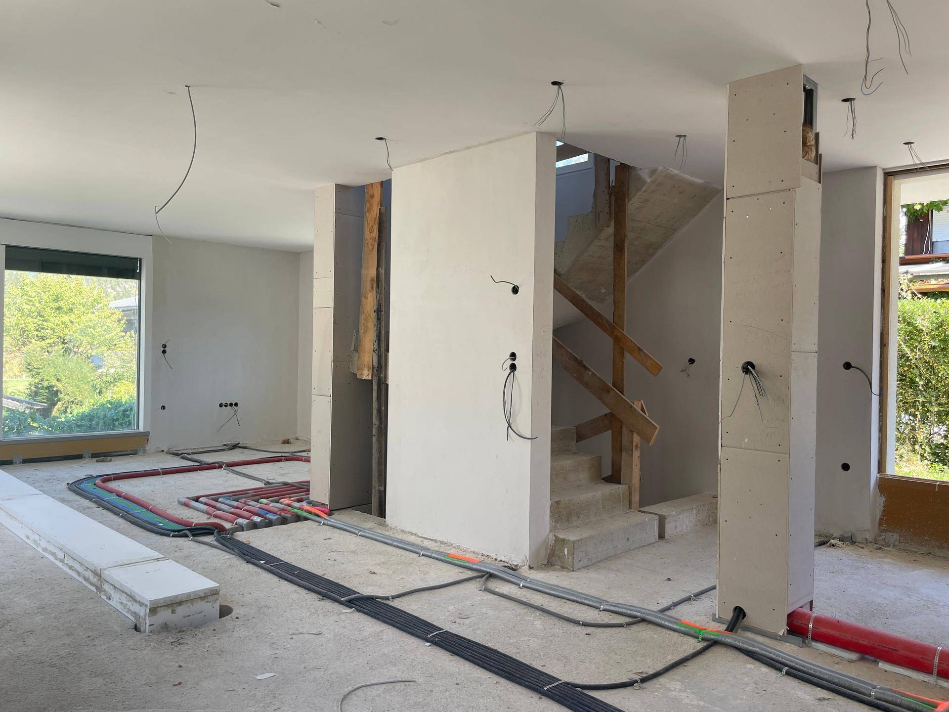 Innenraum im Rohbau mit sichtbaren Installationen, einer Betontreppe und großen Fenstern.