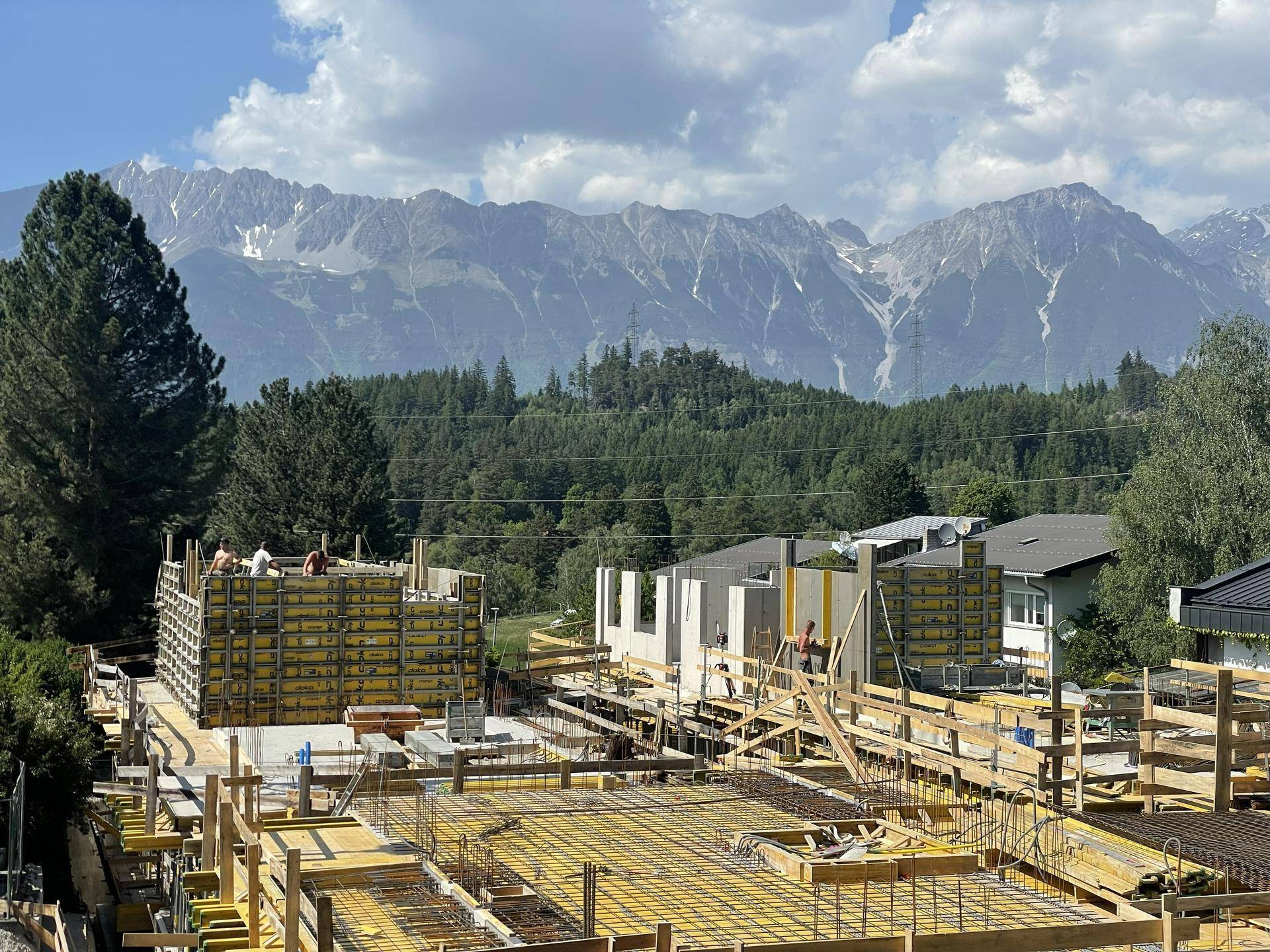 Baustelle mit mehreren im Bau befindlichen Gebäuden vor einer beeindruckenden Bergkulisse und grüner Landschaft.