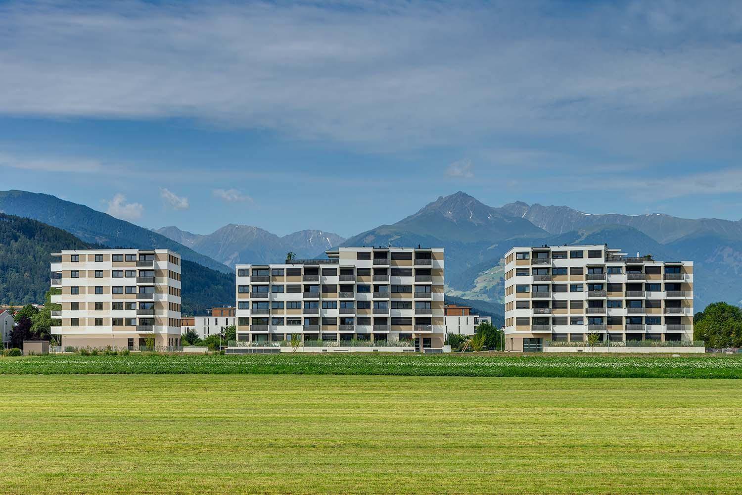 Moderne Wohngebäude mit Balkonen vor einer malerischen Bergkulisse und grünen Feldern.