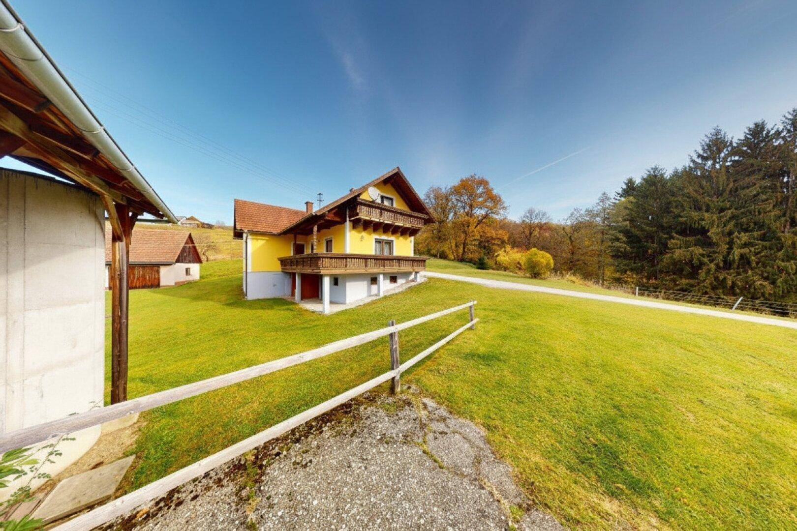 Gelbes Landhaus mit Holzbalkon und rotem Ziegeldach, umgeben von einer weitläufigen grünen Wiese.