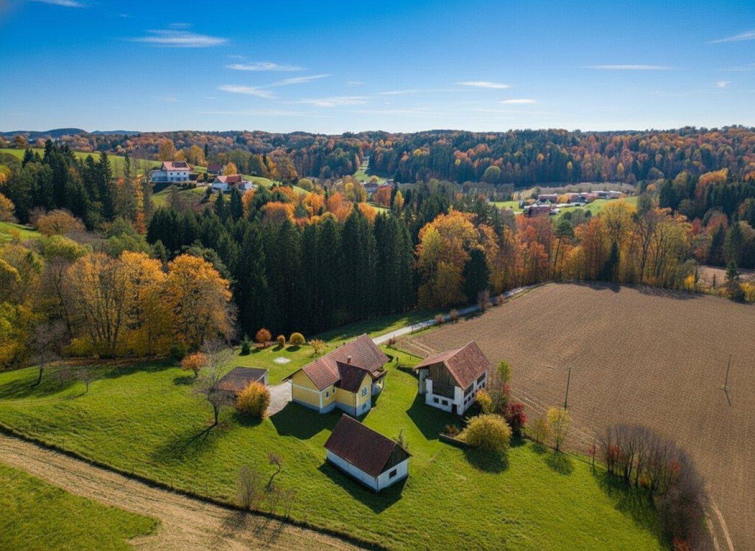 Weitläufige Landschaft mit mehreren Häusern, Feldern und bunten Herbstwäldern unter blauem Himmel.