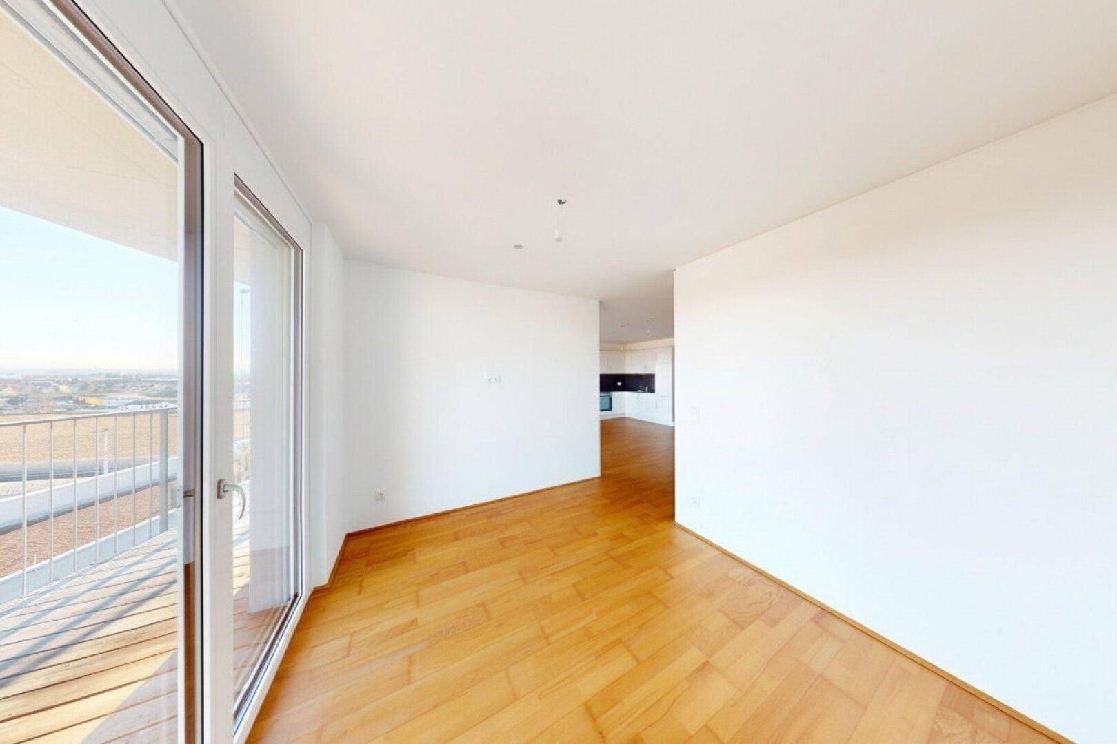 Lichtdurchfluteter Raum mit Holzboden und großem Fenster, das Zugang zu einem Balkon bietet.