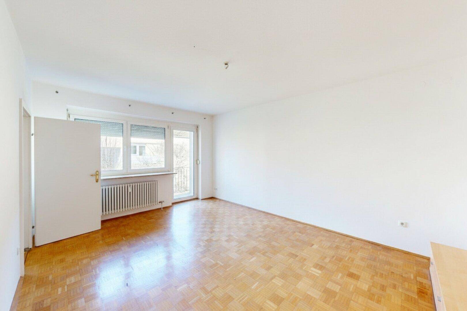 Helles, leeres Wohnzimmer mit Parkettboden und Zugang zu einem kleinen Balkon.