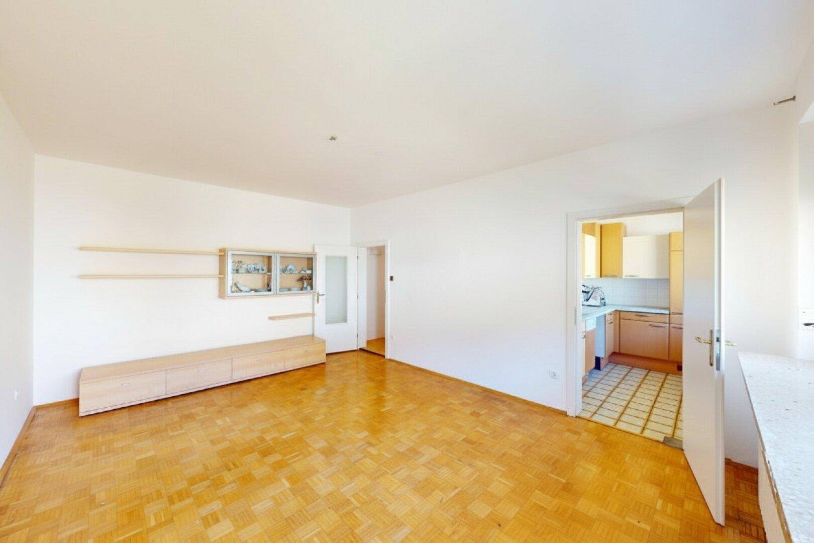 Geräumiges Wohnzimmer mit Parkettboden und offenem Durchgang zur Küche.