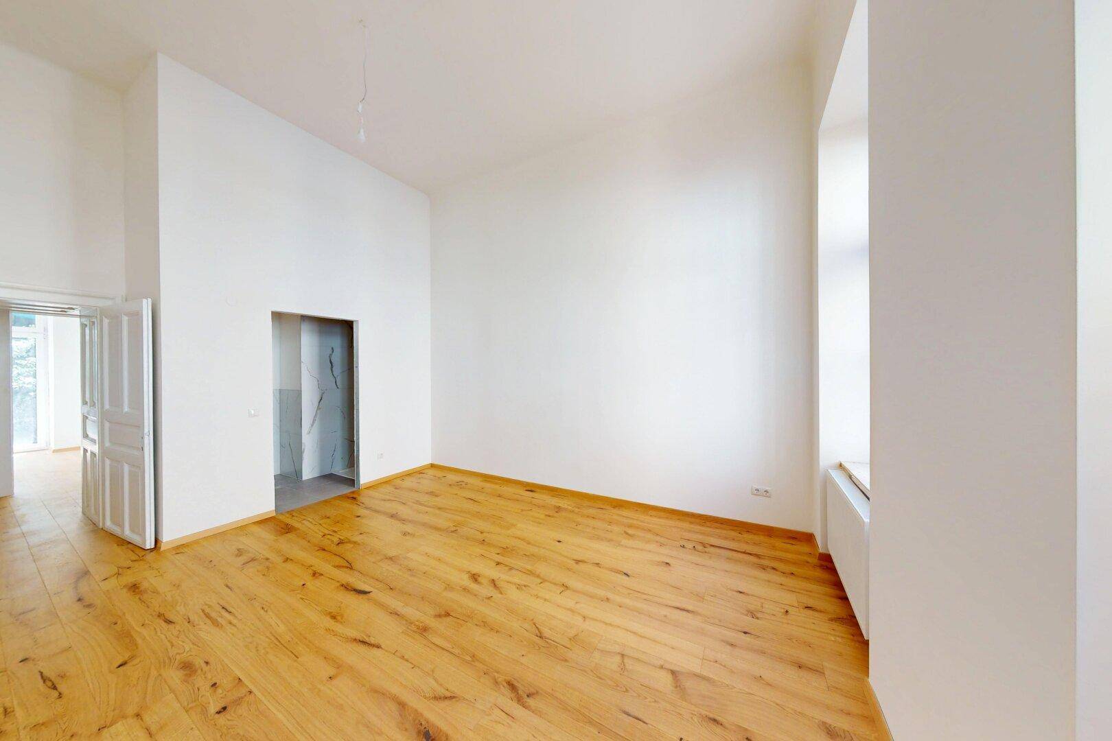 Geräumiger, heller Raum mit Holzboden und Zugang zu einem gefliesten Bereich, möglicherweise ein Bad.