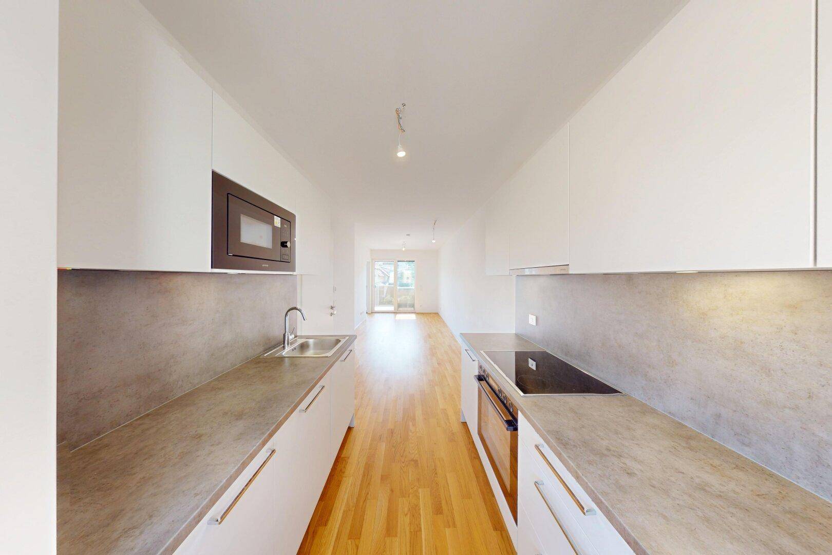 Moderne, schmale Küche mit weißen Schränken, grauen Arbeitsplatten und Holzboden, offen zum Wohnbereich.