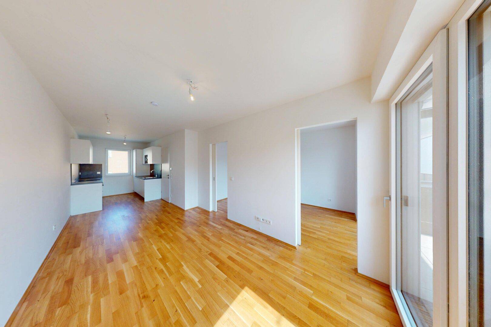 Offener Wohnbereich mit Holzboden und integrierter Küche, bietet Zugang zu einem Balkon und viel Licht.