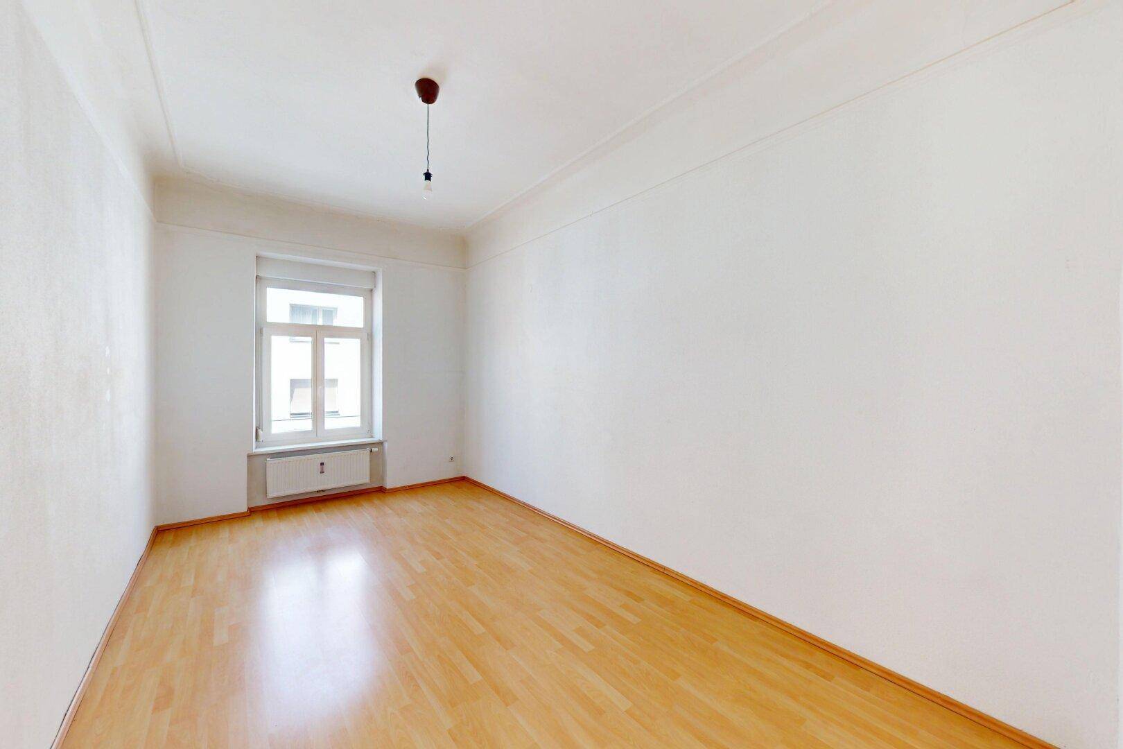 Geräumiges, leeres Zimmer mit Parkettboden, hohen Decken und einem großen Fenster.