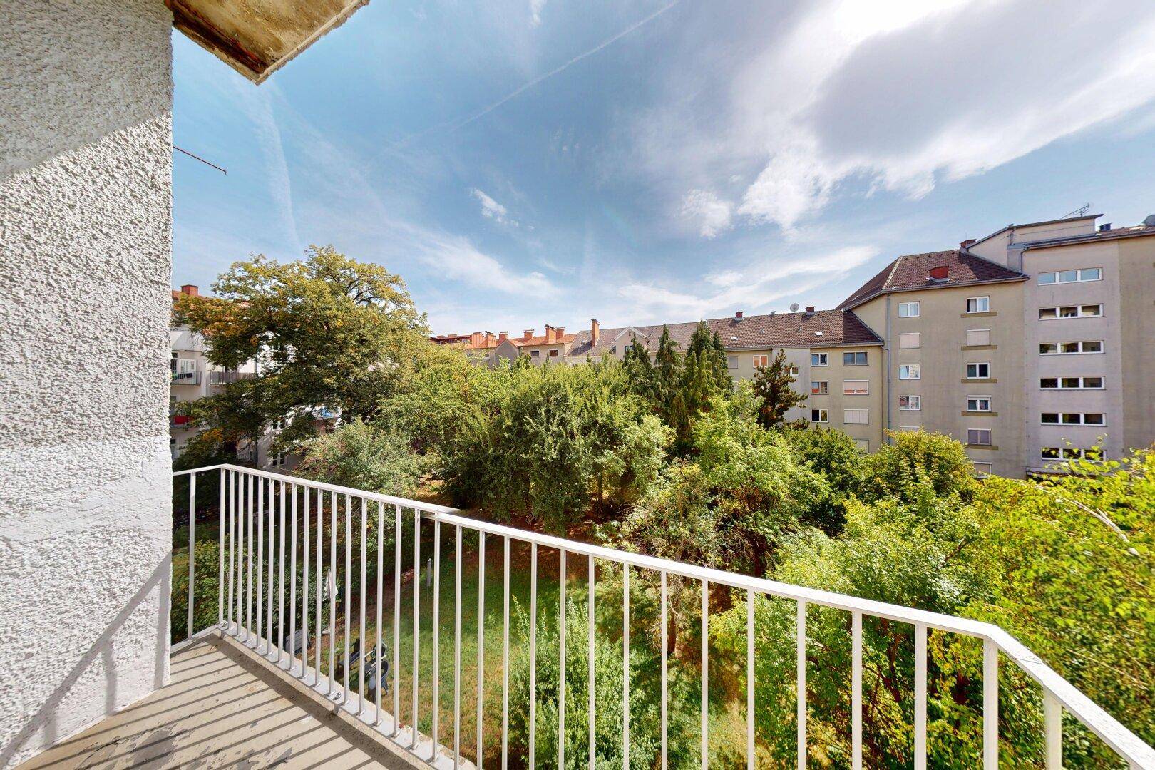 Kleiner Balkon mit Metallgeländer und Blick auf grüne Bäume und umliegende Gebäude.