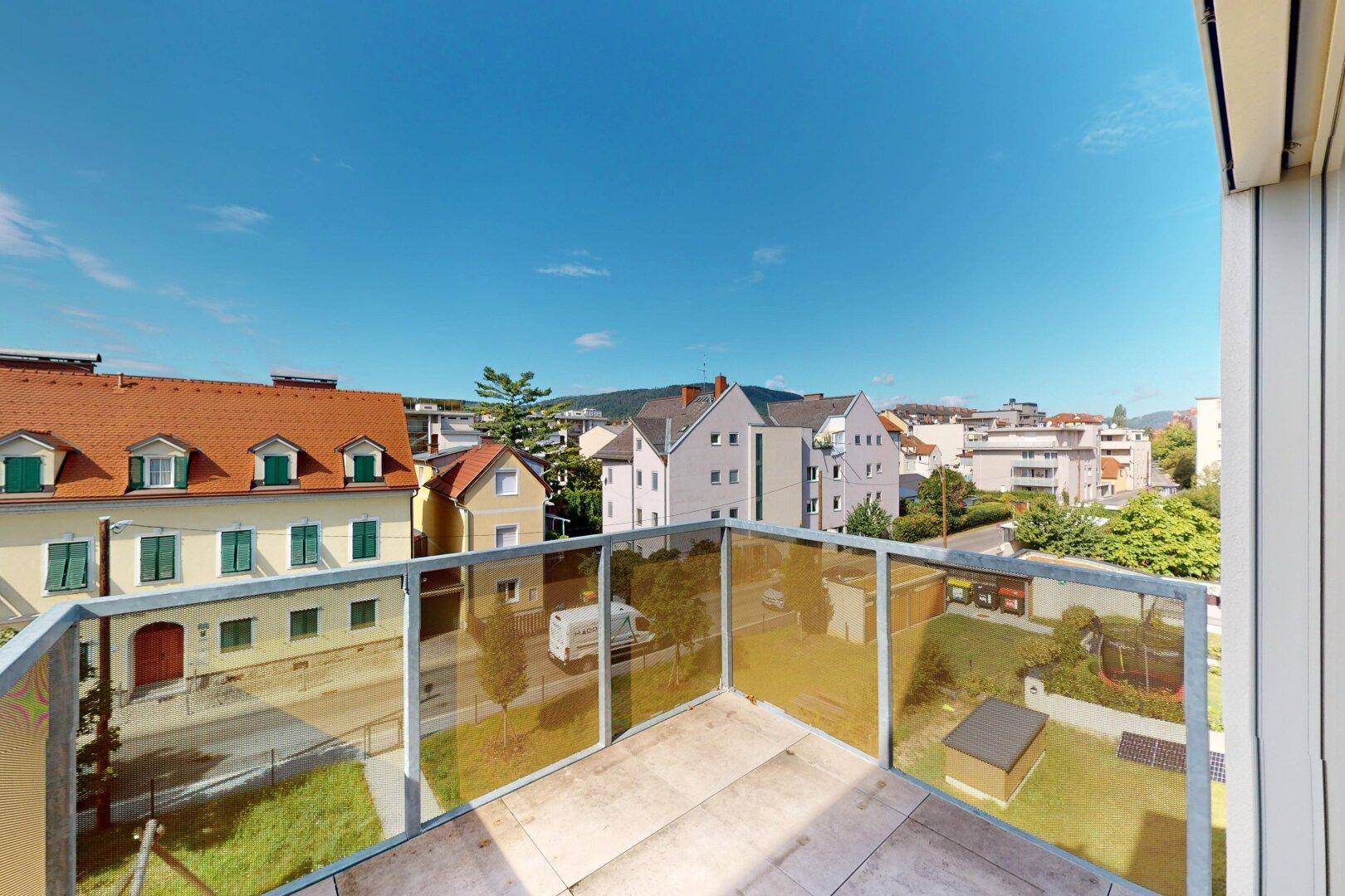 Balkon mit Ausblick auf die umliegende Wohngegend, Gärten und entfernte Hügel unter blauem Himmel.