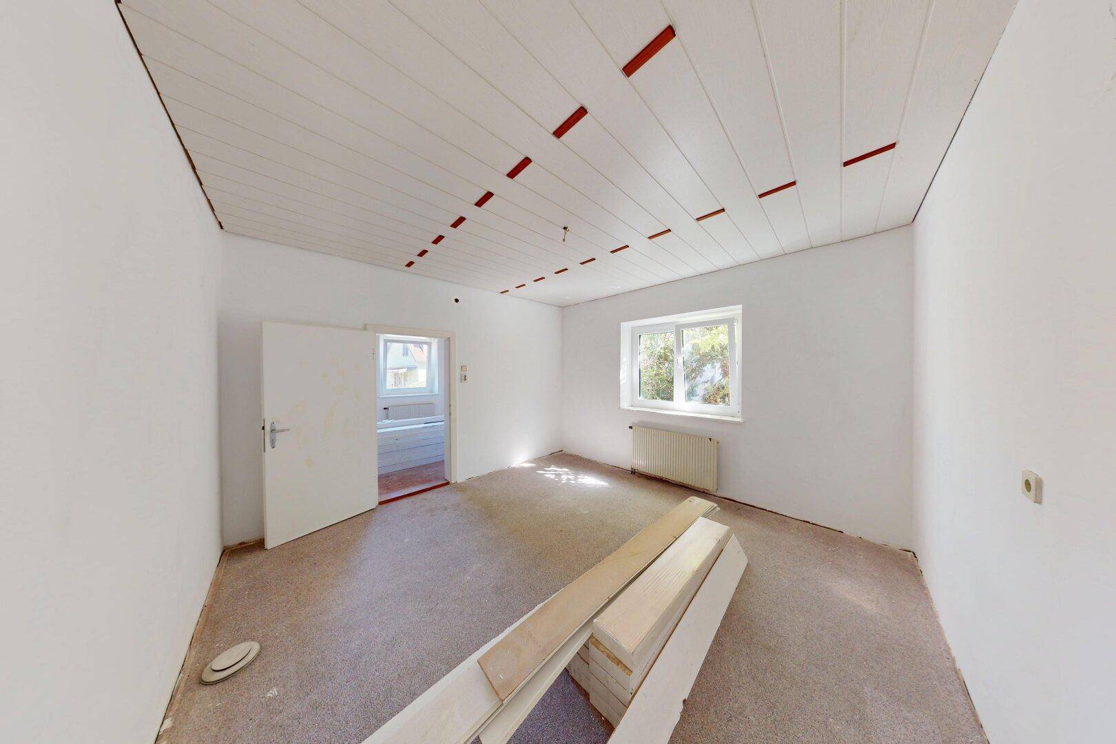 Leerer Raum mit weißer Holzdecke, Fenster und gestapelten neuen Bodenbelägen für die Renovierung.