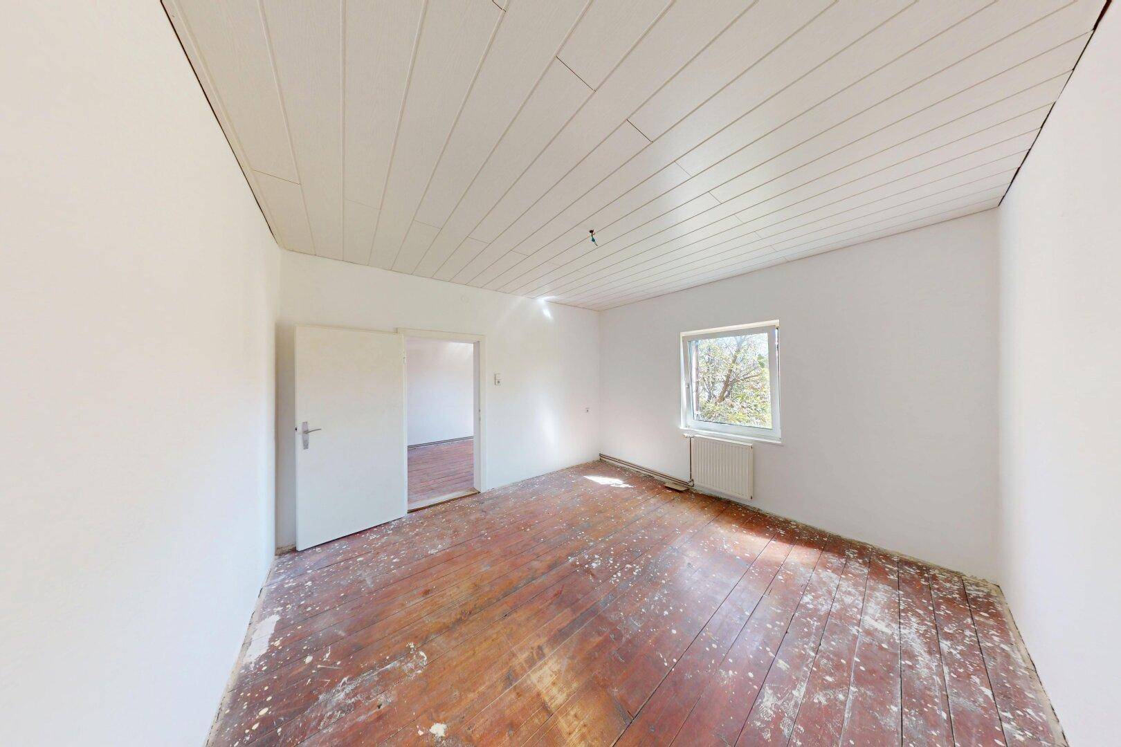 Leerer Raum mit weißer Holzdecke, Fenster und altem Holzdielenboden, bereit zur Renovierung.