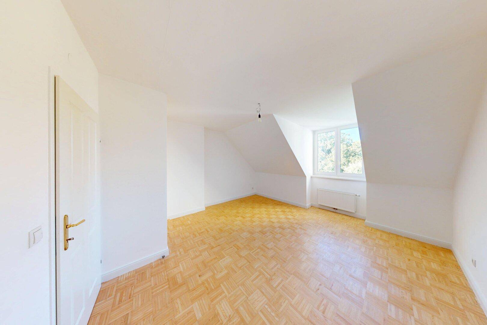 Freundlicher Raum mit Parkettboden, Dachschrägen und einem Fenster, das viel Licht hereinlässt.
